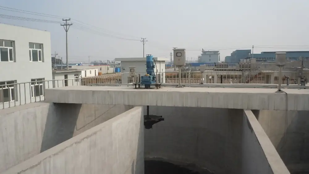 An industrial water treatment facility with large, empty concrete settling tanks, featuring a blue mixing motor unit mounted on a bridge over a circular tank, all set against an outdoor background of factory buildings and power lines