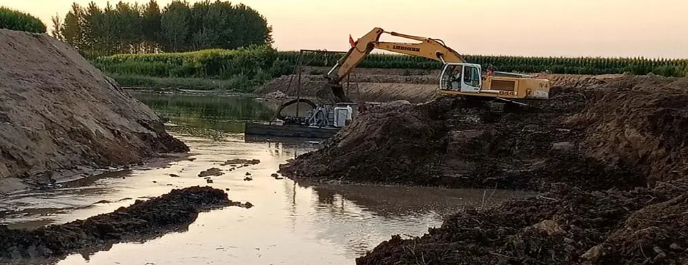 Excavator dredging a muddy water channel for sediment removal